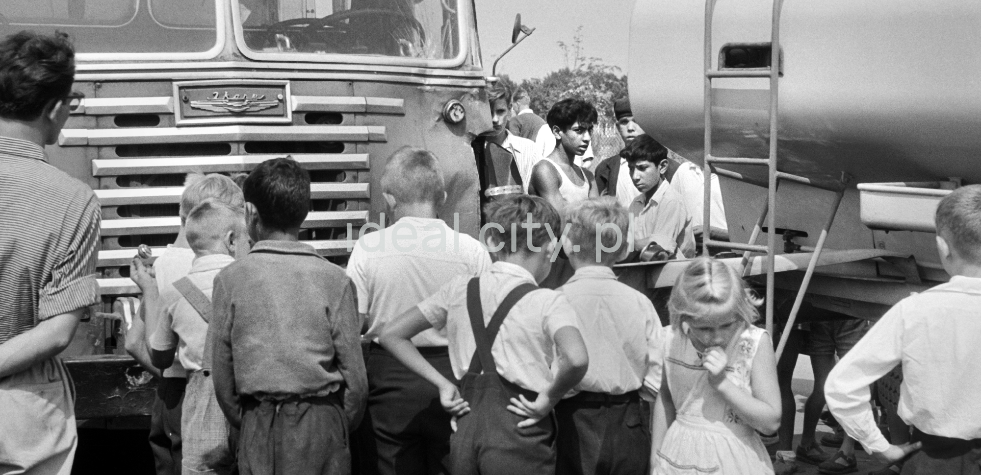 A group of children between the head of the bus and the cistern in front of it.
