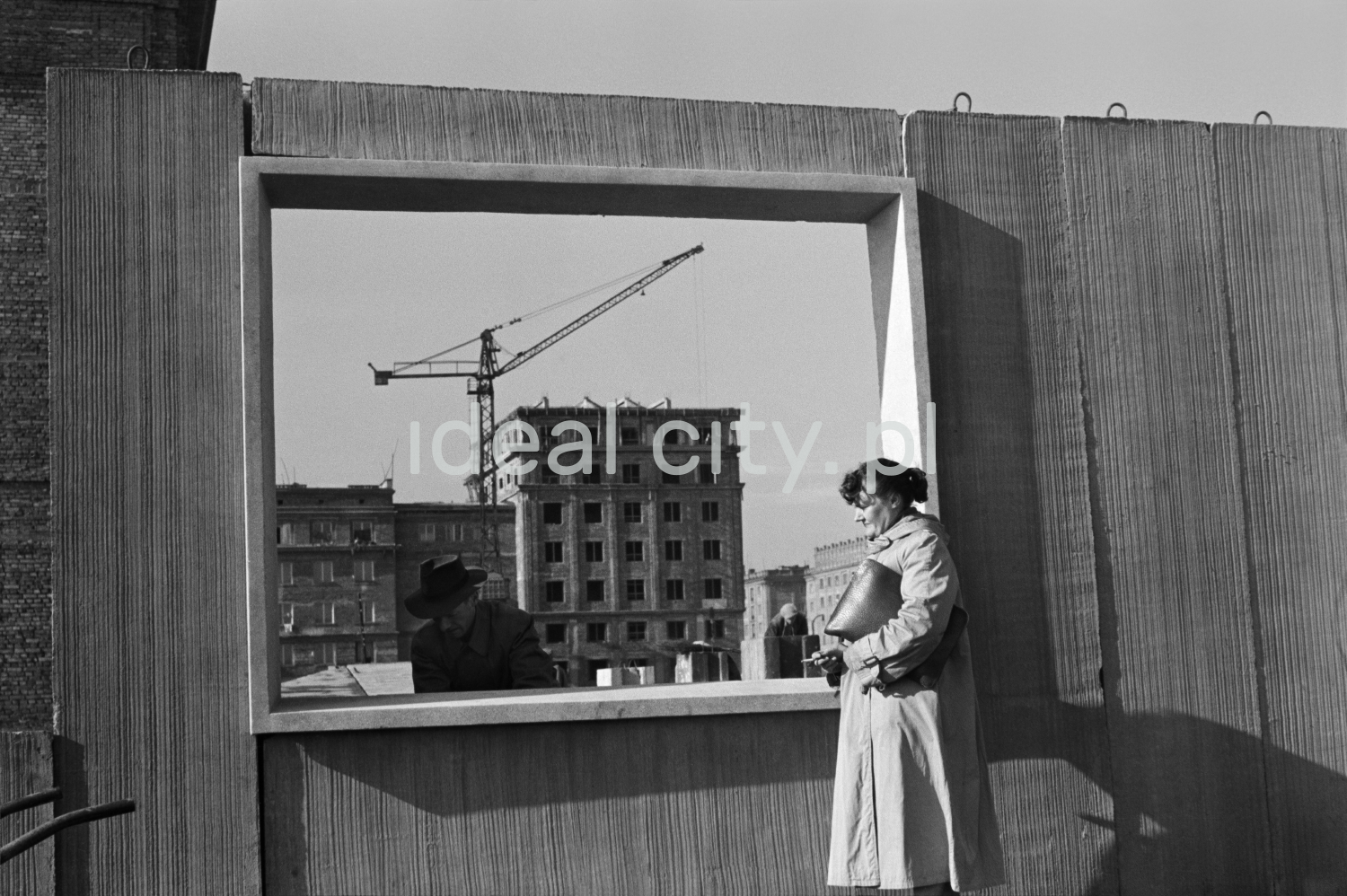 A woman in a trench coat and a briefcase under her arm stands against a prefabricated wall with a window opening.