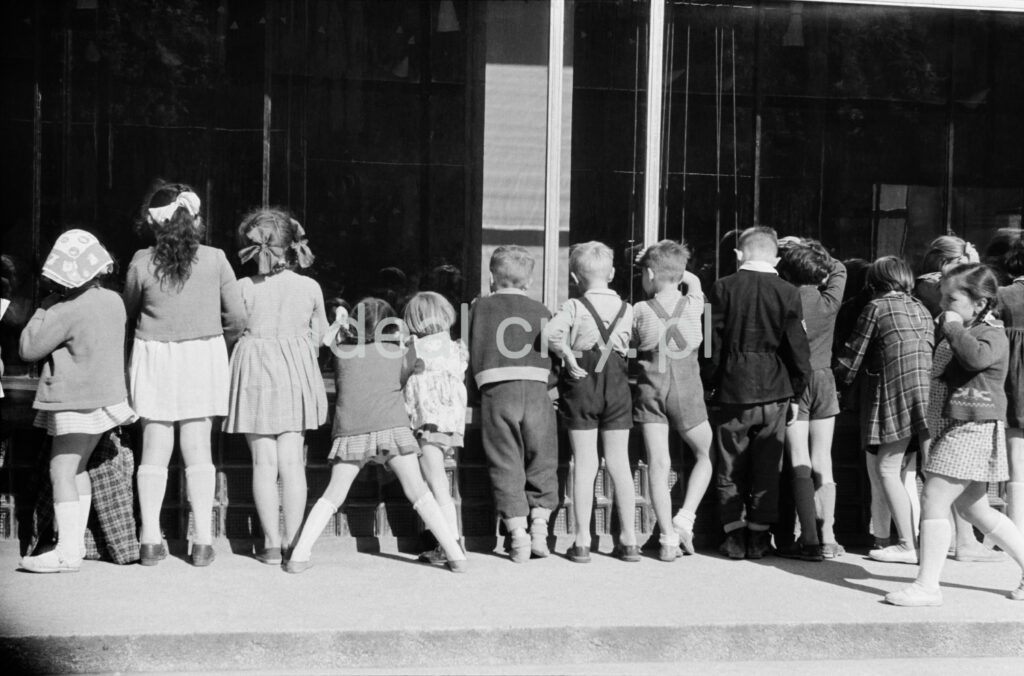 A group of children and several women look through the window into the interior of a shop on the ground floor of a modernist building.