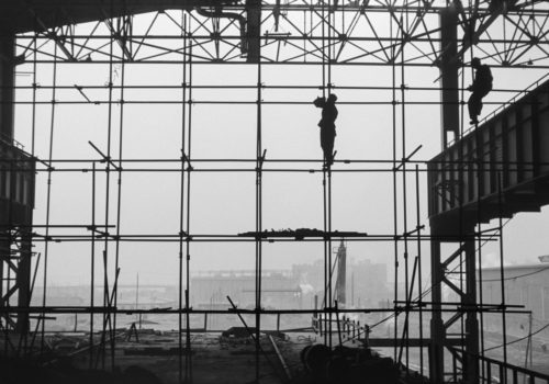 Dwóch mężczyzn w robotniczych strojach balansuje na rusztowaniu, w trakcie budowy ściany zewnętrznej hali fabrycznej. Ujęcie kontrastowe z jej wnętrza z widokiem na pozostałe obiekty na terenie kombinatu.