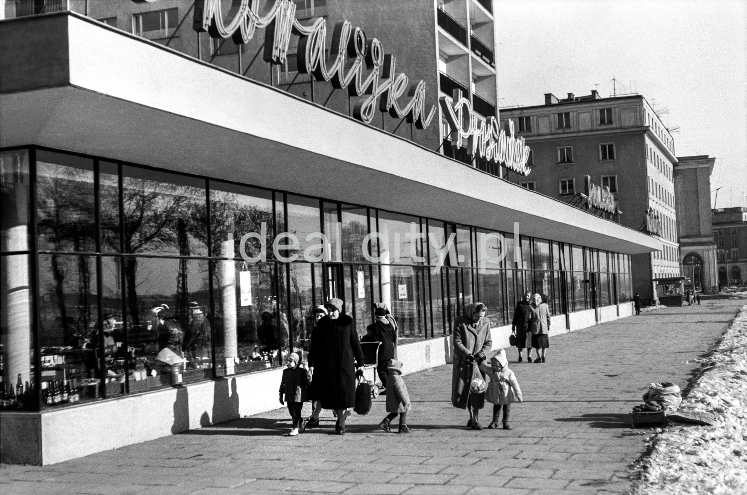 Pedestrian traffic on a wide sidewalk along the line of shop pavilions located on the ground floor of a high block of flats.