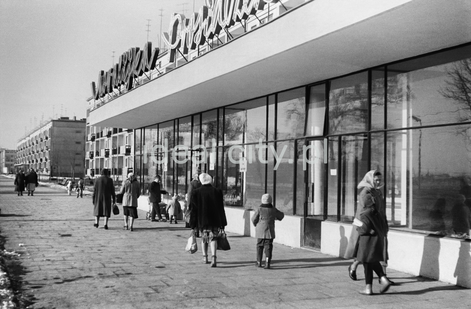 Pedestrian traffic on a wide sidewalk along the line of shop pavilions located on the ground floor of a high block of flats.
