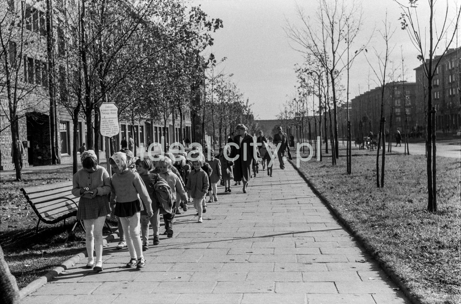 Children in school clothes march along the sidewalk that stretches along the green belt with residential buildings behind it.