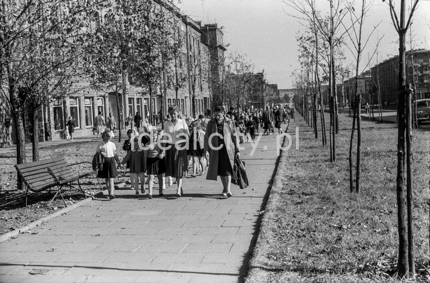 Children in school clothes are marching along the sidewalk that stretches along the green belt and residential buildings behind it, accompanied by two teachers at the front.