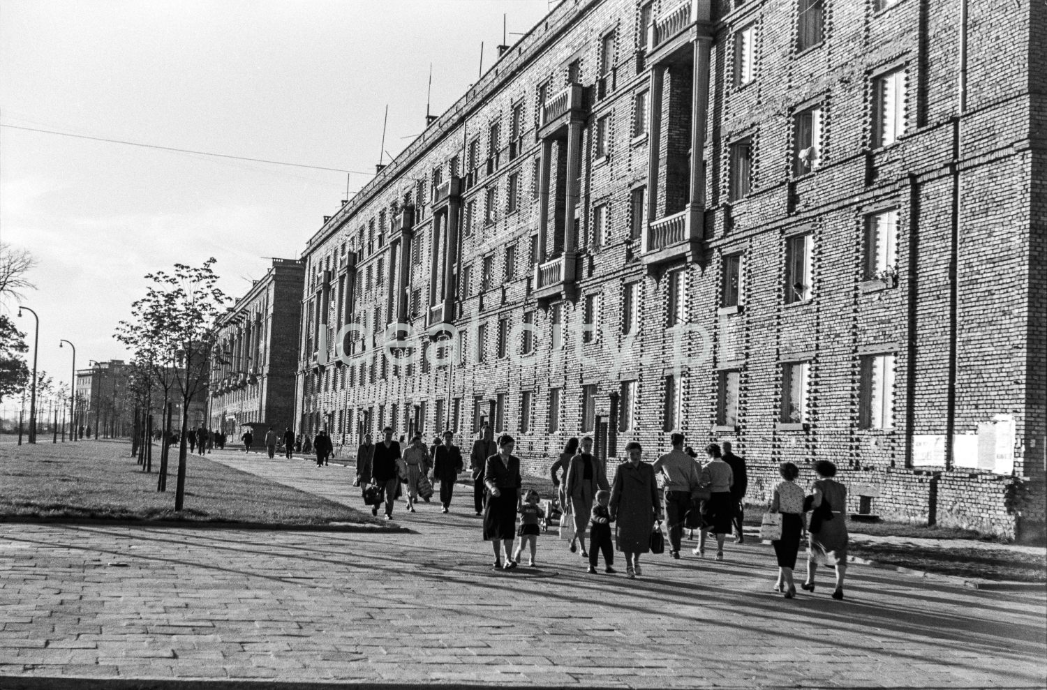 Pedestrian traffic on a wide pavement along an unplastered brick blocks.