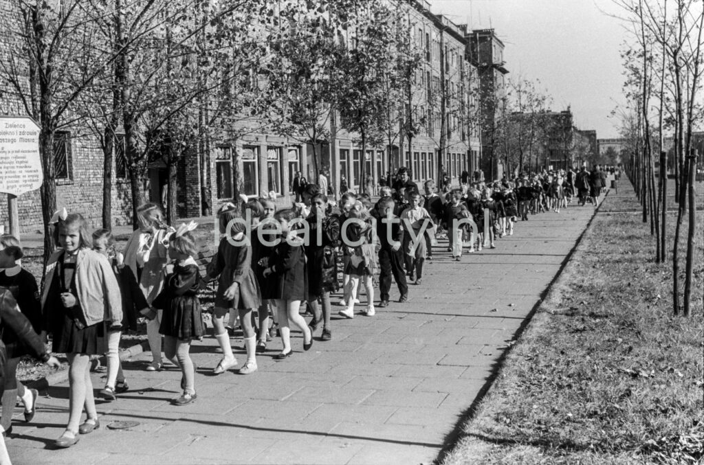 Children in school clothes march along the sidewalk that stretches along the green belt with residential buildings behind it.