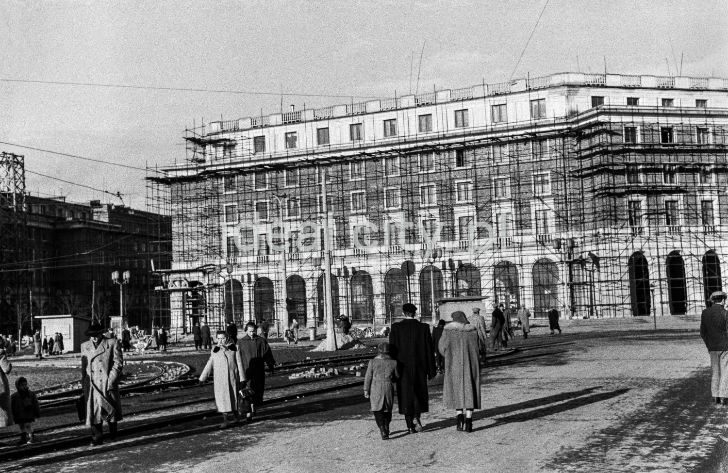 Pedestrian traffic in a spacious city square, in the background scaffolding on monumental residential buildings inspired by the Renaissance.