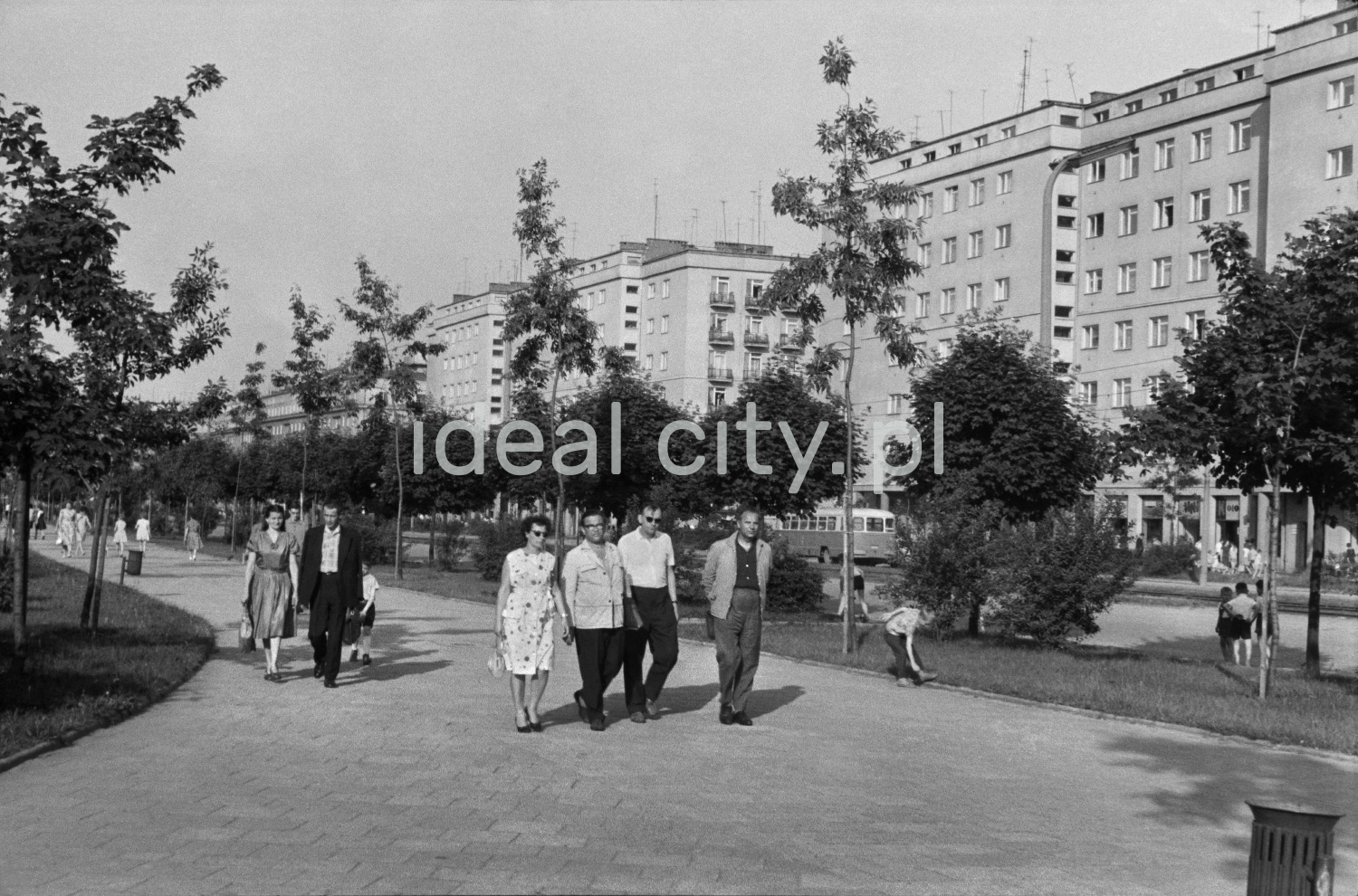 Front shot of pedestrians walking along a wide sidewalk running through a green area, in the background the monumental buildings of the block.