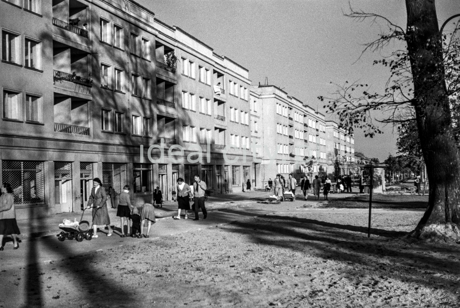Pedestrian traffic on a wide pavement along a row of low apartment blocks, a tree on the right.