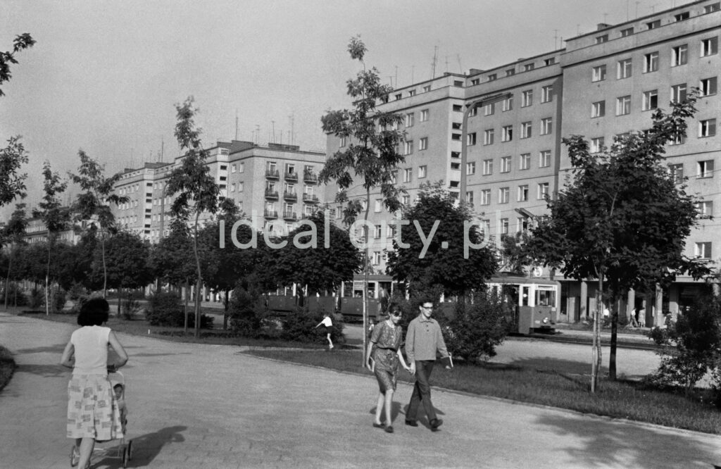 Pedestrian traffic on the wide pavement running through the green area, in the foreground a couple of young people. A massive apartment block in the background.