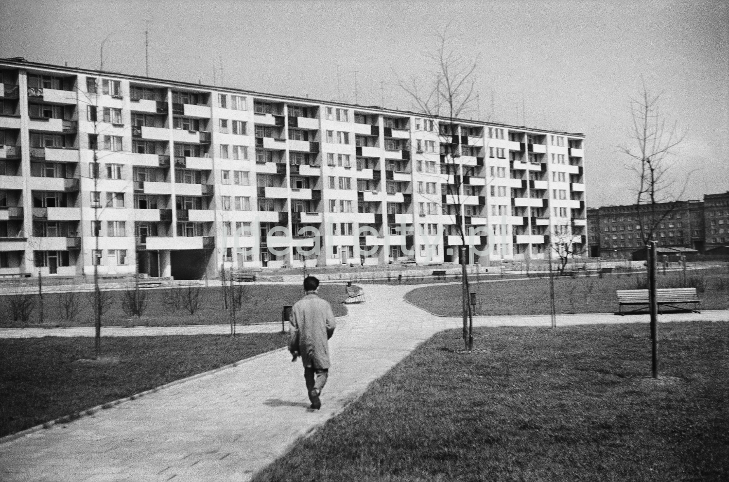 A man in a trench coat walks dynamically along the pavement towards a wide, modernist block of flats.