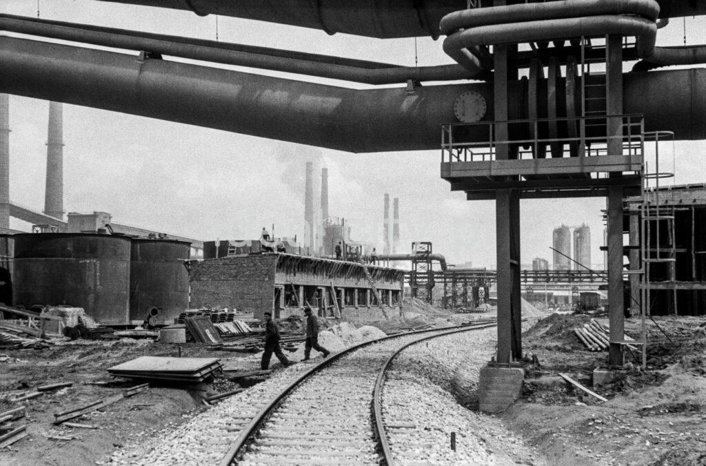 Two men cross the railroad tracks between the infrastructure of the plant.