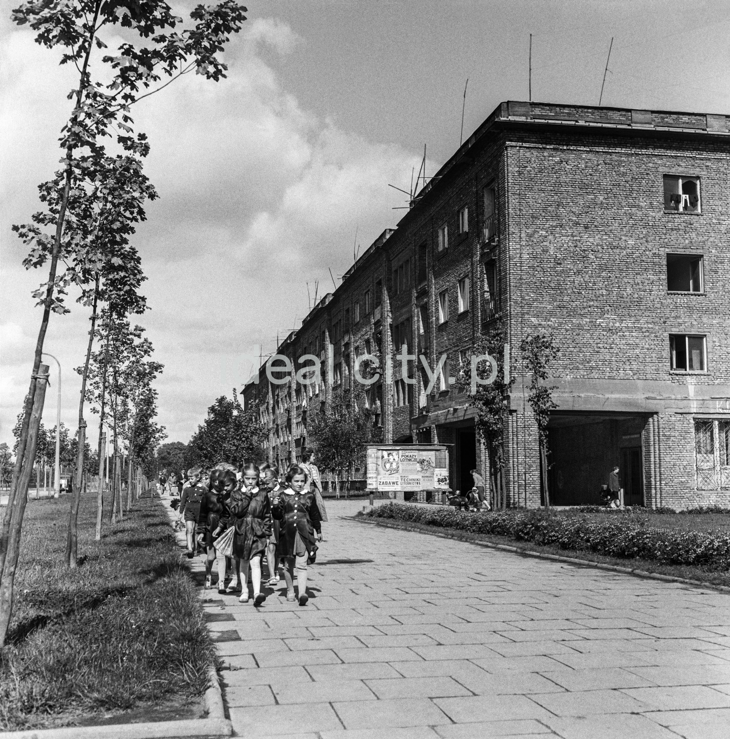 Children in school uniforms march along the paved sidewalk, on the right an unplastered brick apartment block.