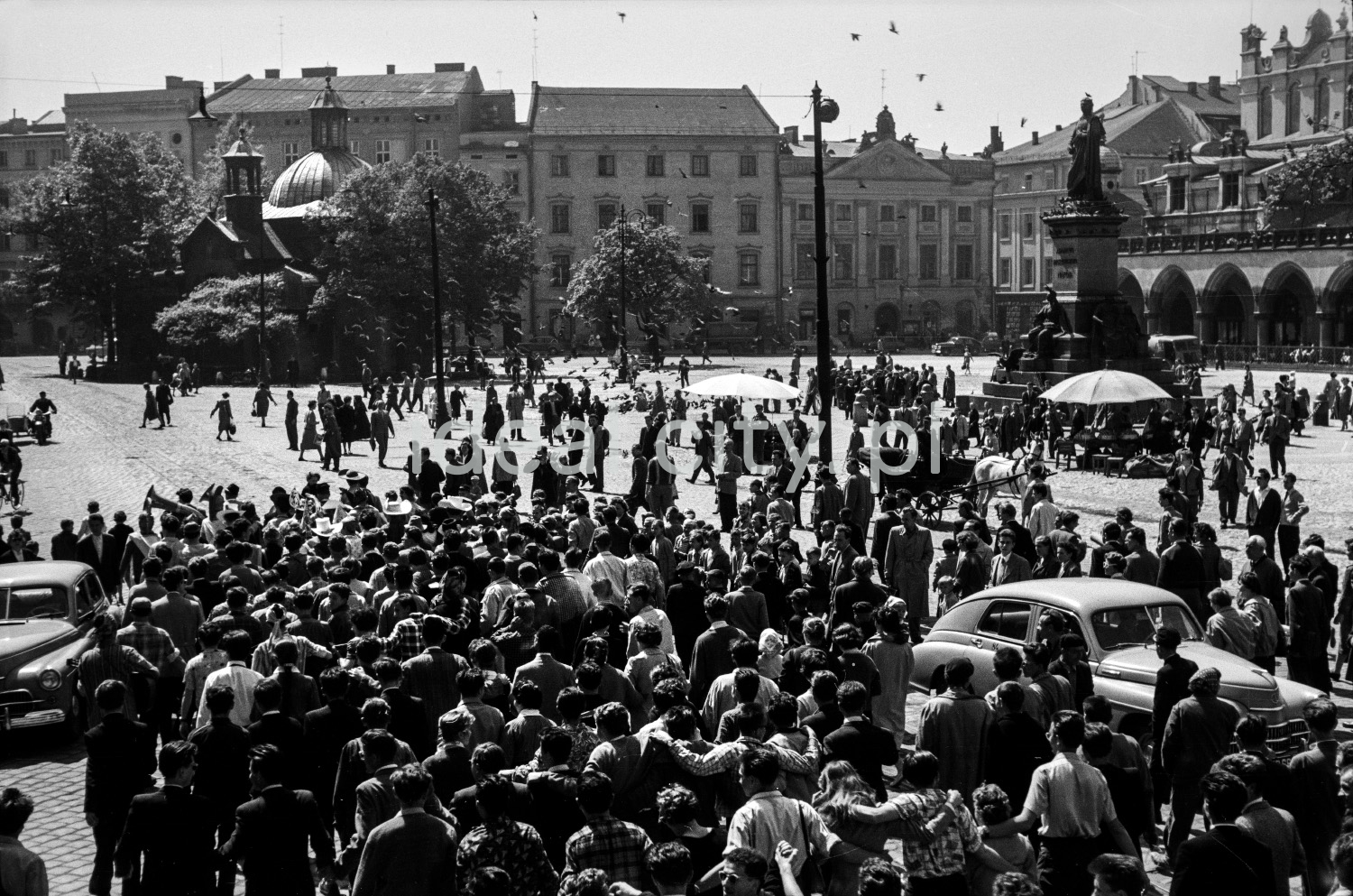 A crowd of people in the town square surrounded by Renaissance tenement houses, a monument and a small church in the background.