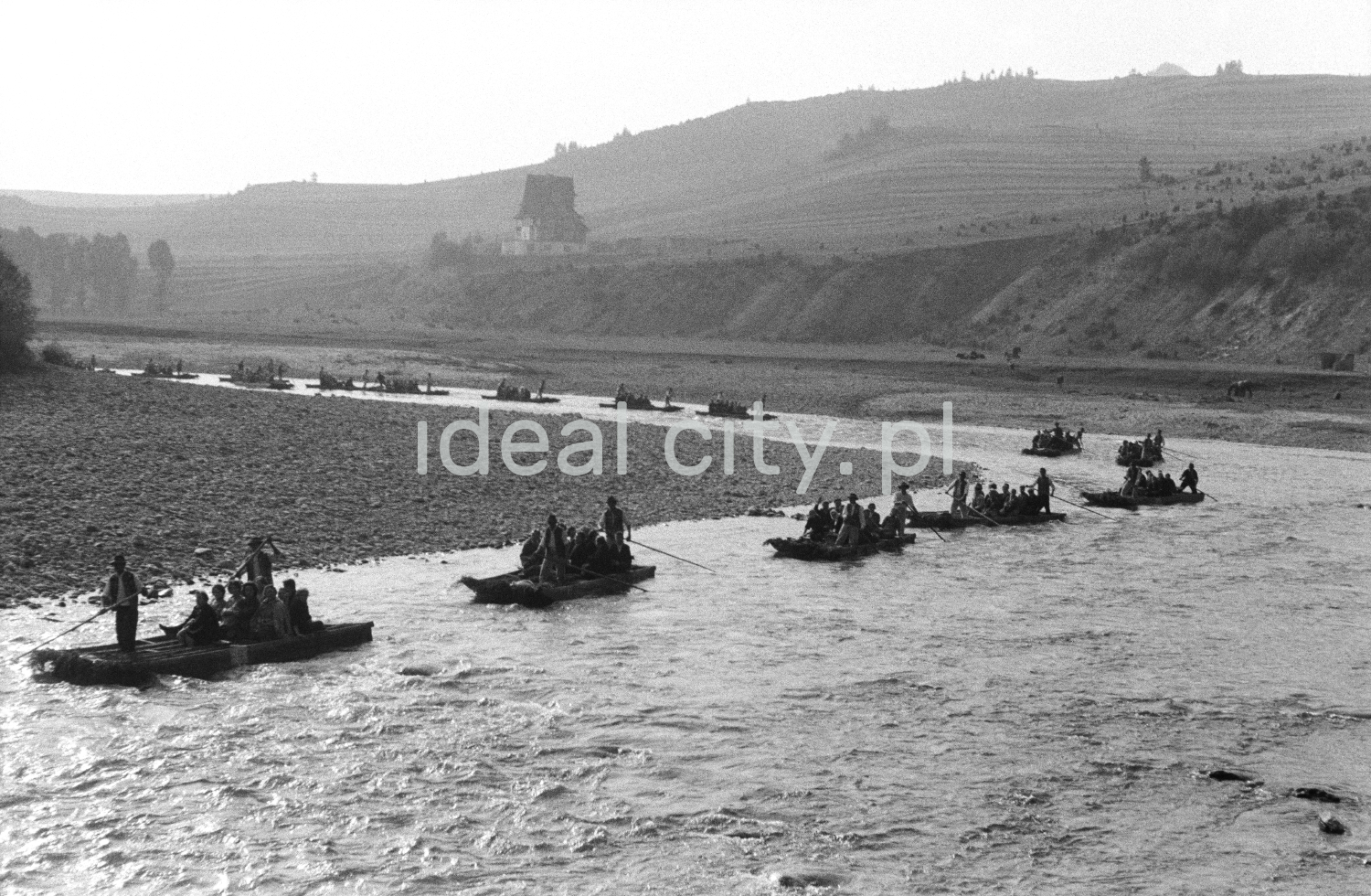 A line of rafts with tourists led by raftsmen flows along a bend in a wide, shallow river.