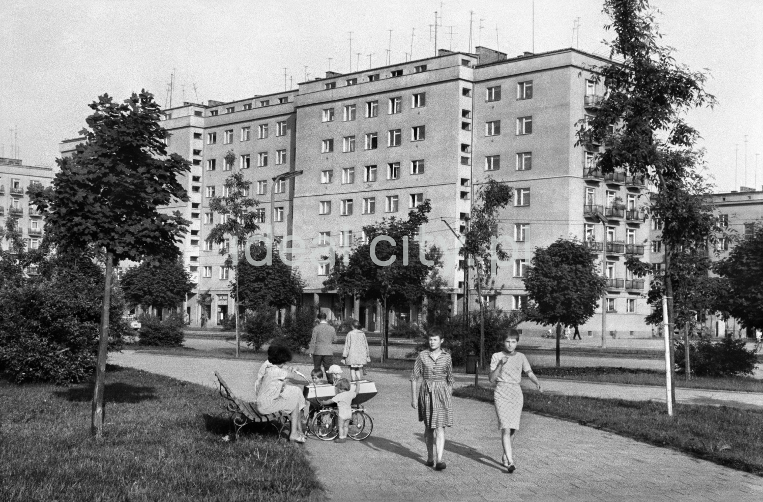 Pedestrian traffic on the wide pavement running through the green area, in the foreground a couple of young people. A massive apartment block in the background.