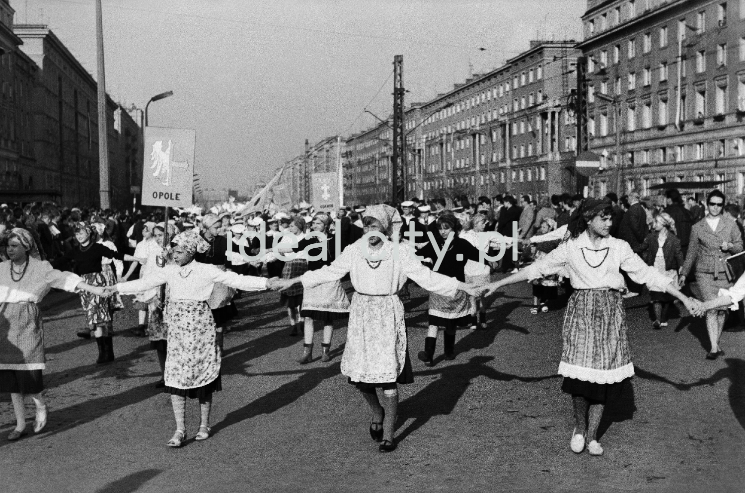 Girls in national costumes walk down a wide alley holding hands.