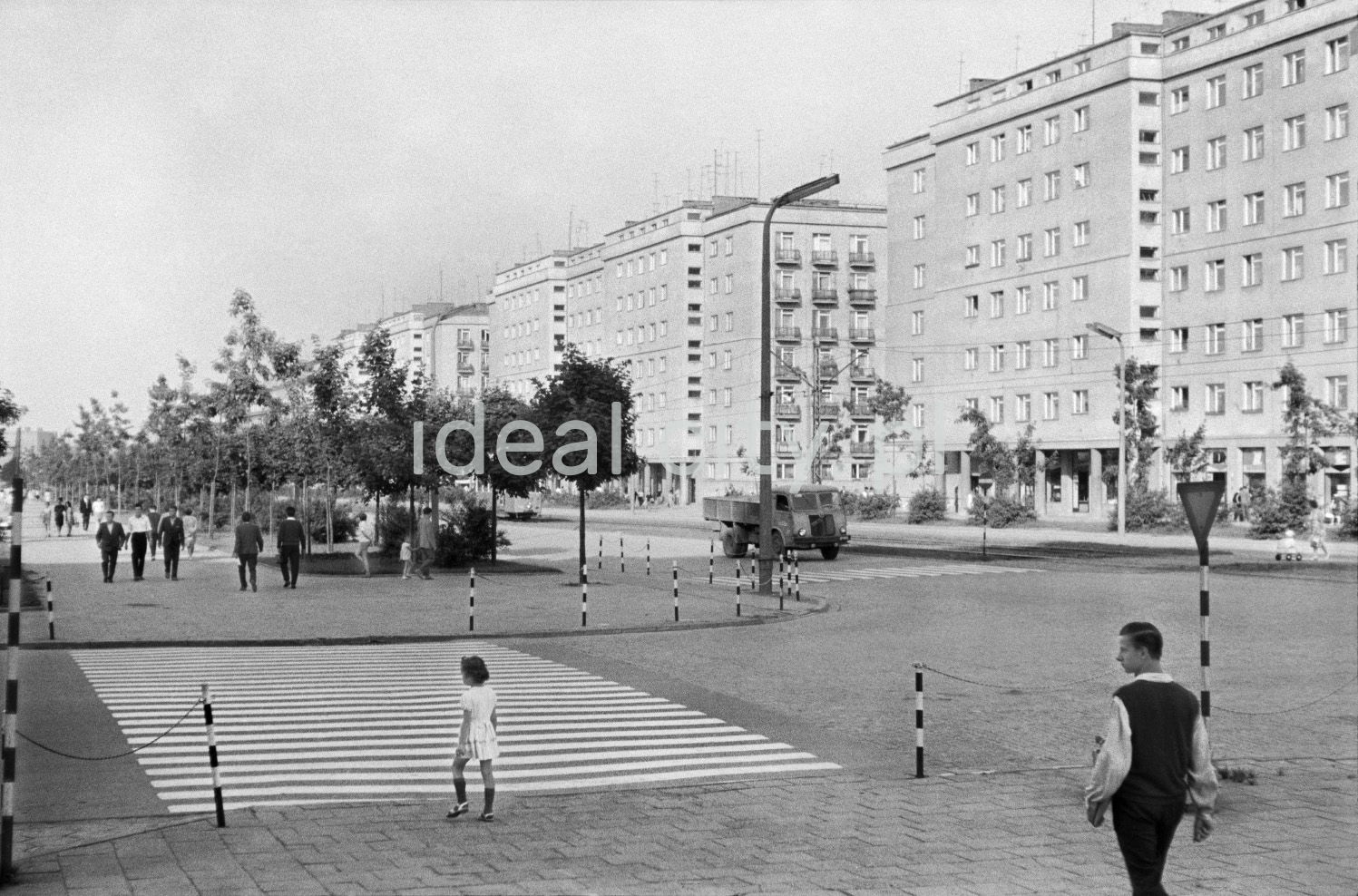 A girl in a white dress prepares to cross the wide pedestrian crossing, on the right a series of apartment blocks.