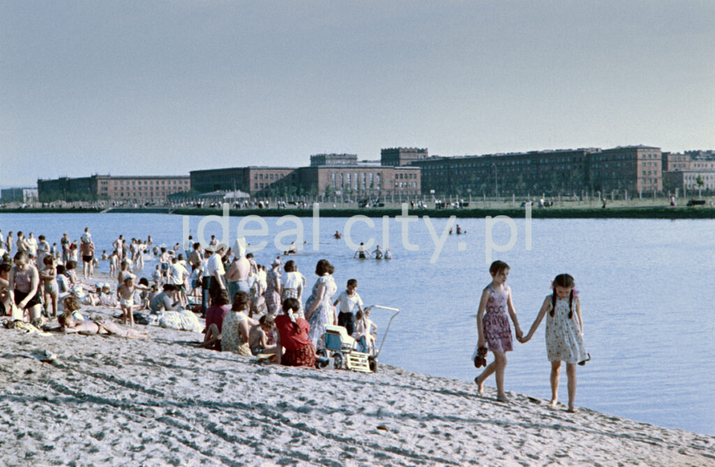 Two girls are walking hand in hand on the sandy beach, then a crowd of people is resting, on the other shore, in the distance, you can see monumental residential buildings.