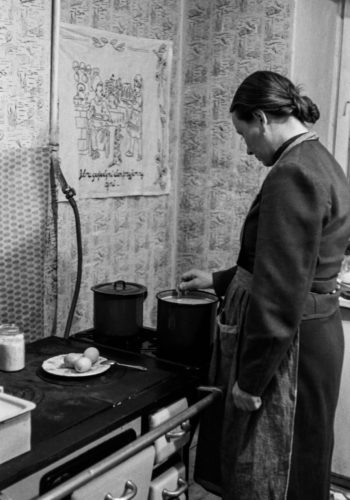 Kobieta w fartuchu przygotowuje posiłek przy kuchni węglowej. Na blacie garnki, talerz z jajkami.