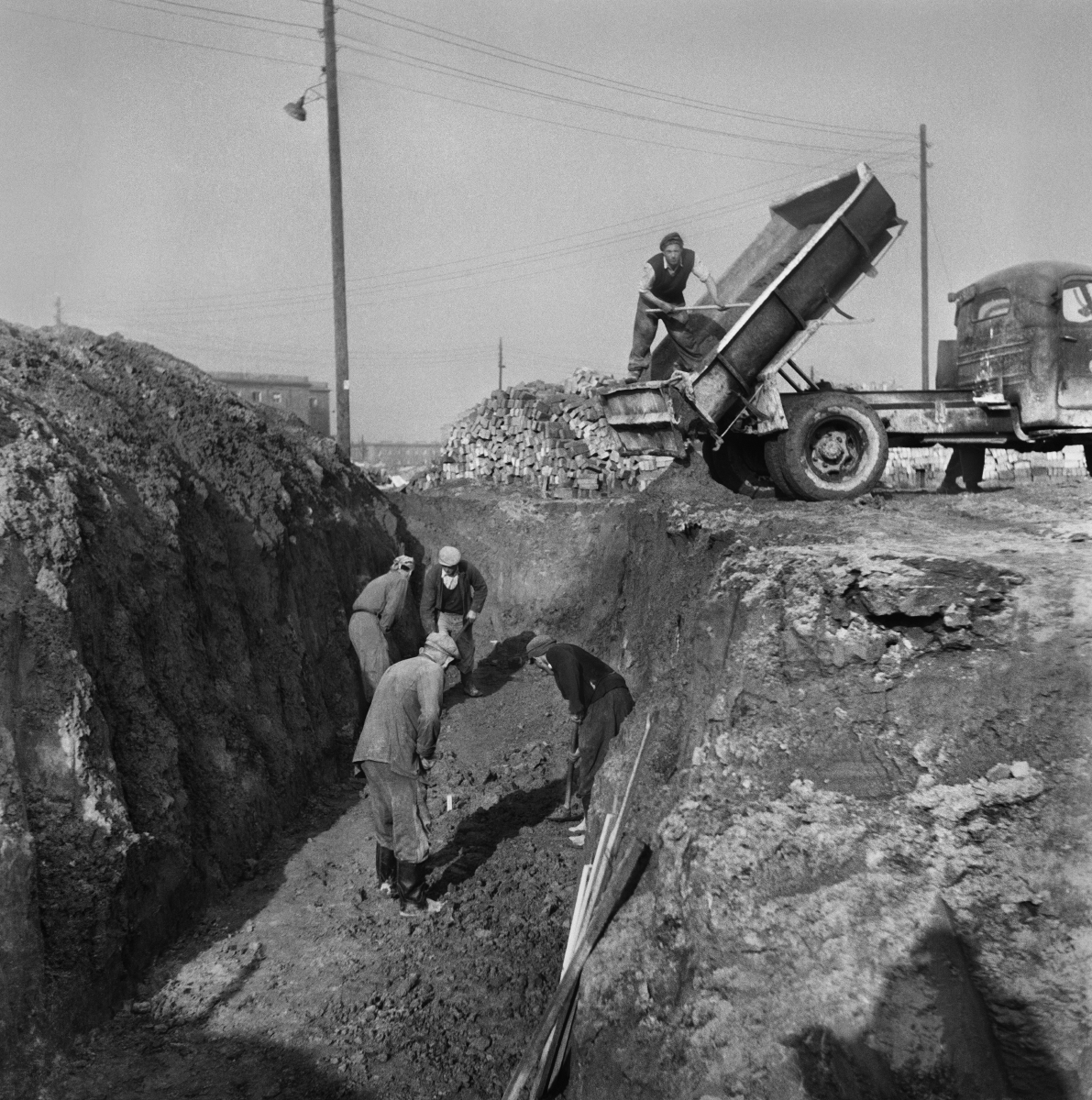 Workers work with shovels in the trench, over them is a dump truck.
