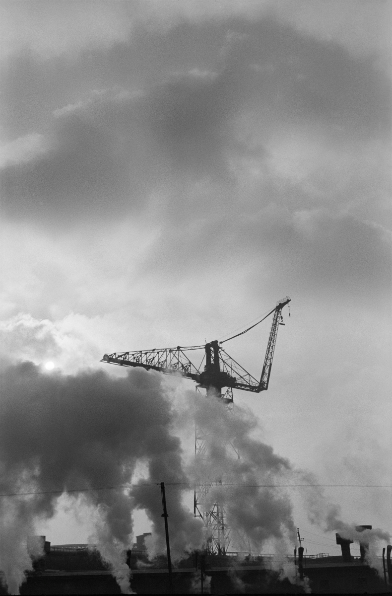 A shot of a construction crane hidden among the clouds of smoke from factory chimneys.