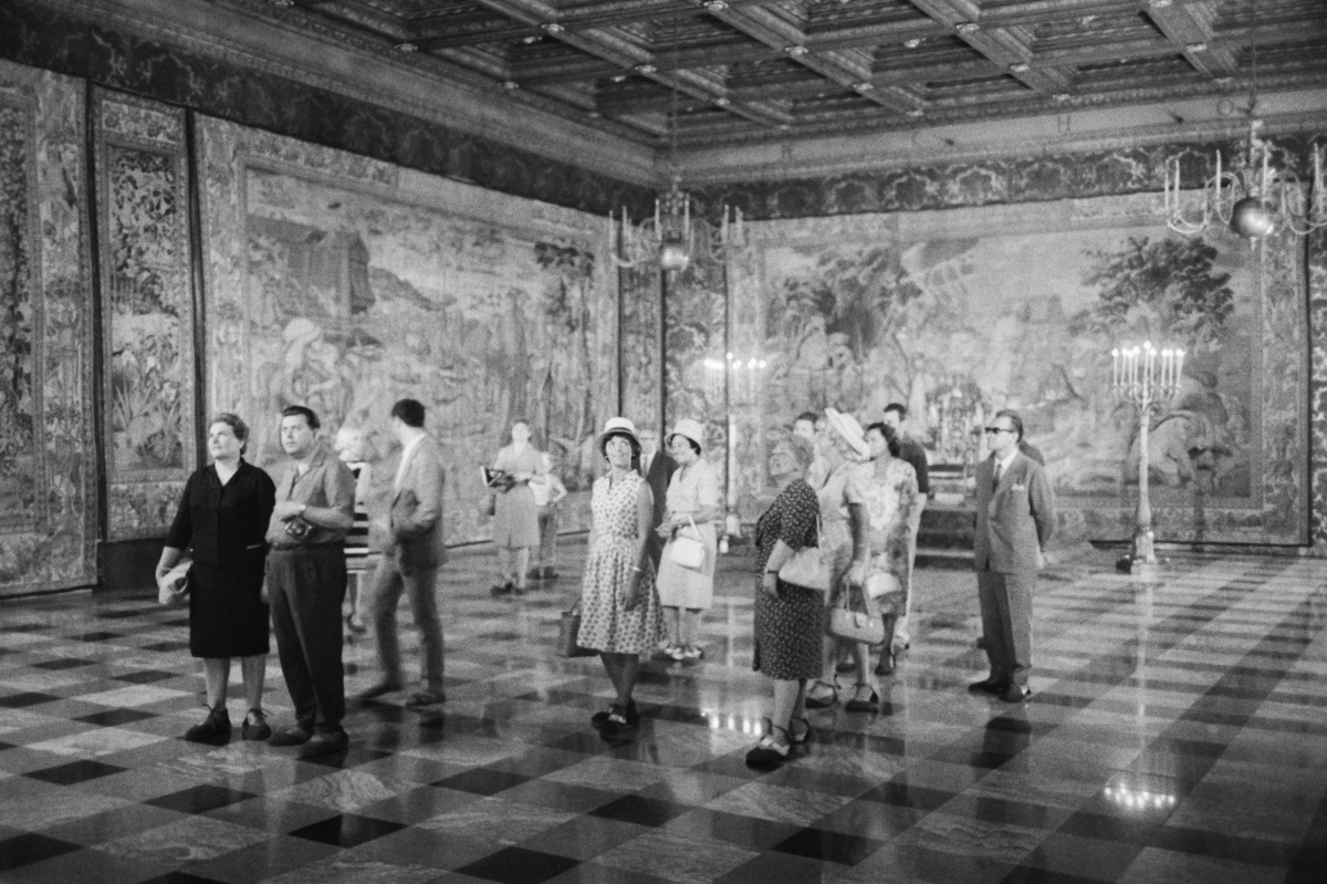 A group of tourists is examining the tapestries on the walls of a spacious room.