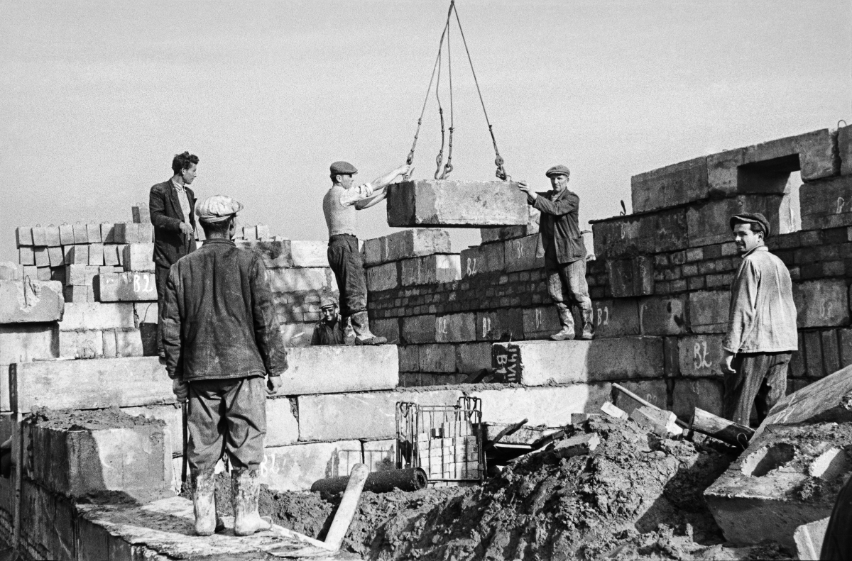 Two workers steer a concrete block fed from above by a crane, a few others watch it.