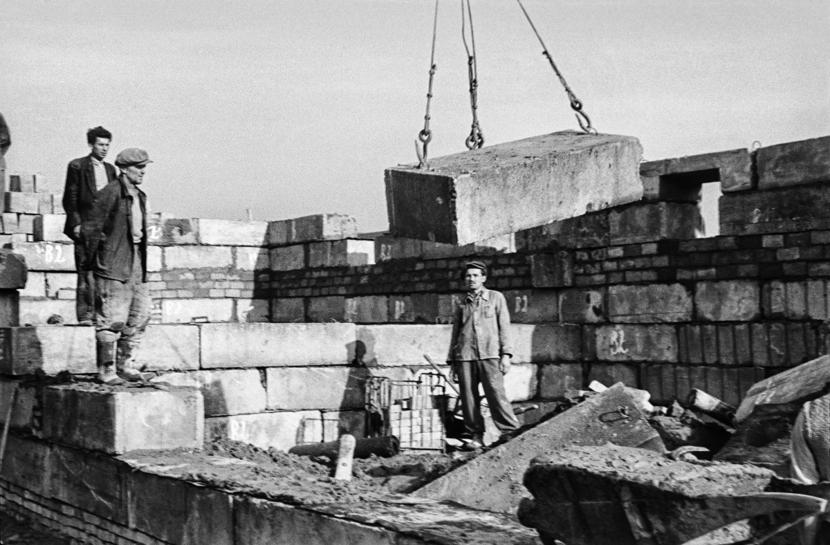 A crane hands over a concrete block and several workers are watching it.