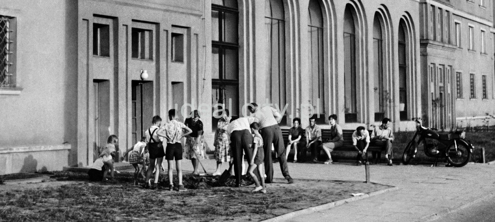 Grupa mieszkańców w trakcie prac społecznych na trawniku przed budynkiem mieszkalnym.
