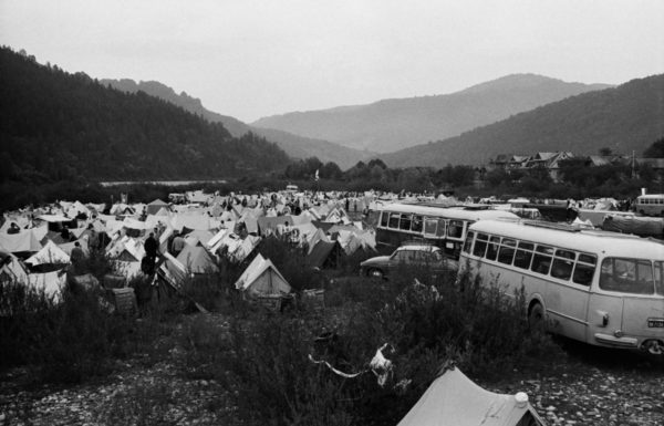 Namiotowe obozowisko w dolinie nad rzeką, pomiędzy namiotami zaparkowane autobusy i auta osobowe.