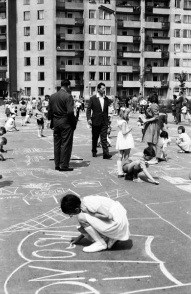 Grupa dzieci pod nadzorem dorosłych rysuje kredą na asfalcie. W tle modernistyczne bloki.