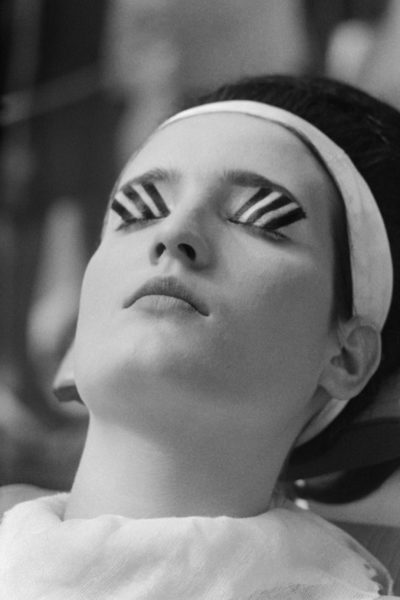 Modelka w trakcie nakładania makijażu op-art. Na zamkniętych powiekach wyraziste pionowe paski, ujęcie z profilu.