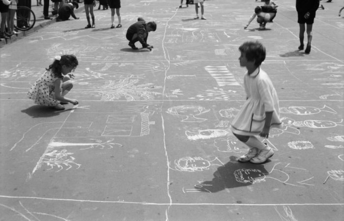 Grupa dzieci pod nadzorem dorosłych rysuje kredą na asfalcie.