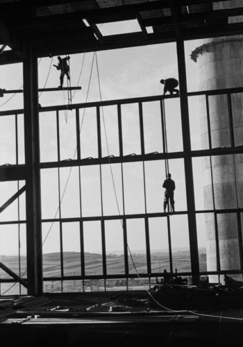 Postaci trzech robotników montują ażurową konstrukcję hali fabrycznej, za nią widoczny pagórkowaty krajobraz.