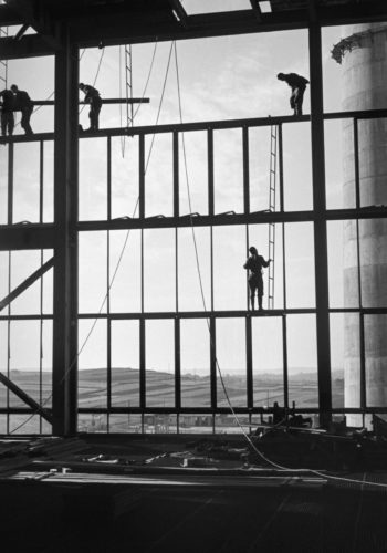 Postaci trzech robotników montują ażurową konstrukcję hali fabrycznej, za nią widoczny pagórkowaty krajobraz.