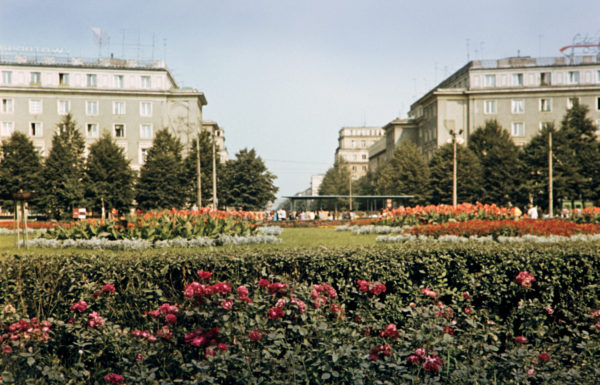Kolorowa fotografia w konwencji pocztówkowej przedstawia pokryty przystrzyżonym trawnikiem plac miejski, klomby z kwiatami, na drugim planie przejeżdża błękitny tramwaj, w tle socrealistyczna zabudowa zwieńczona neonem "Składamy oszczędności w PKO"