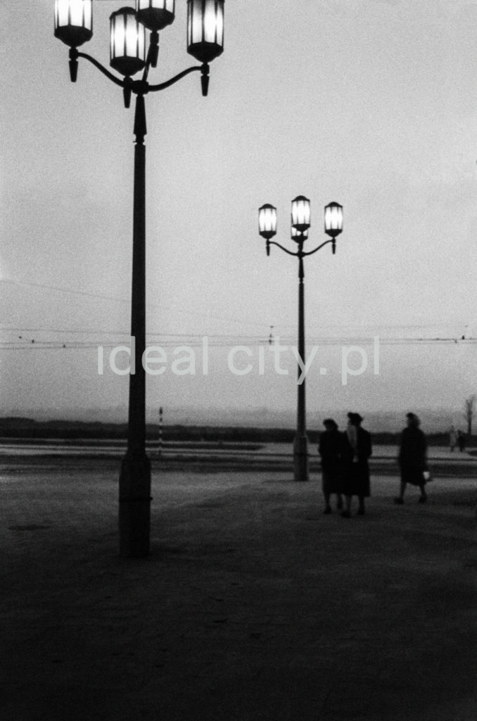 Pionowe ujęcie na szereg rozświetlonych latarni ulicznych w stylu socrealistycznym, na chodniku sylwetki ludzi.