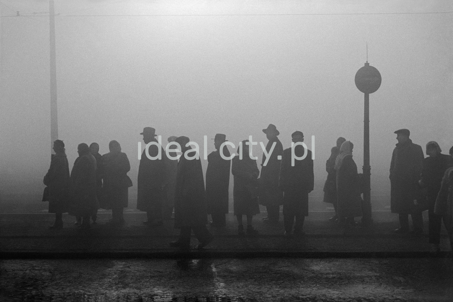 Sylwetki ludzi w płaszczach i kapeluszach stojących we mgle mna przystanku tramwajowym.