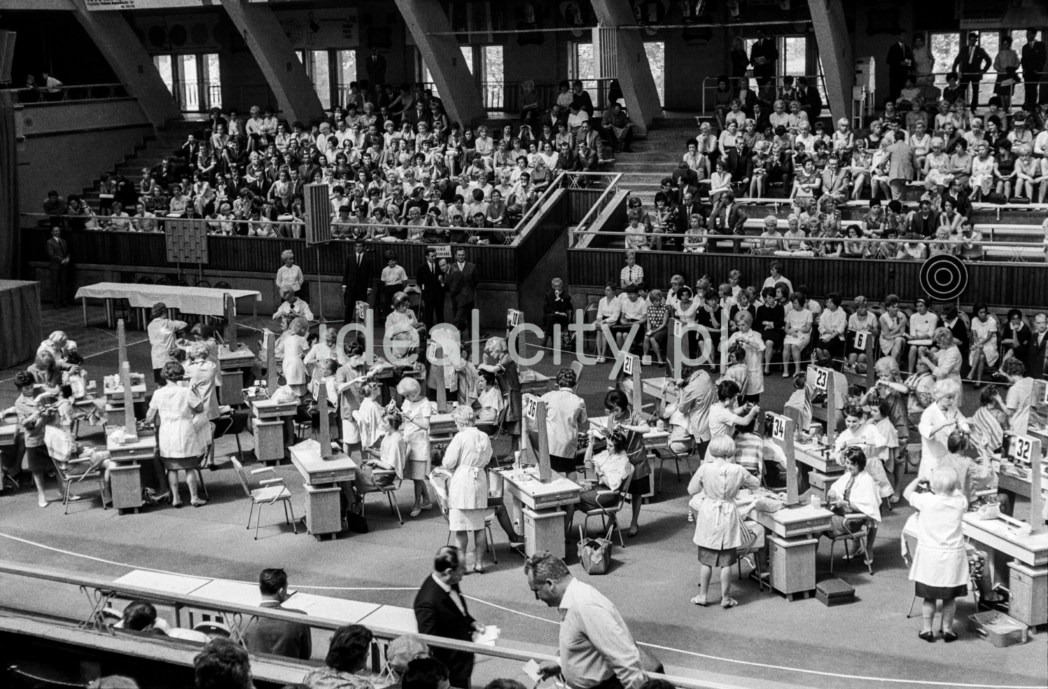 A shot from a height on the tables with participants set up in the center of the hall, and the audience in the background.
