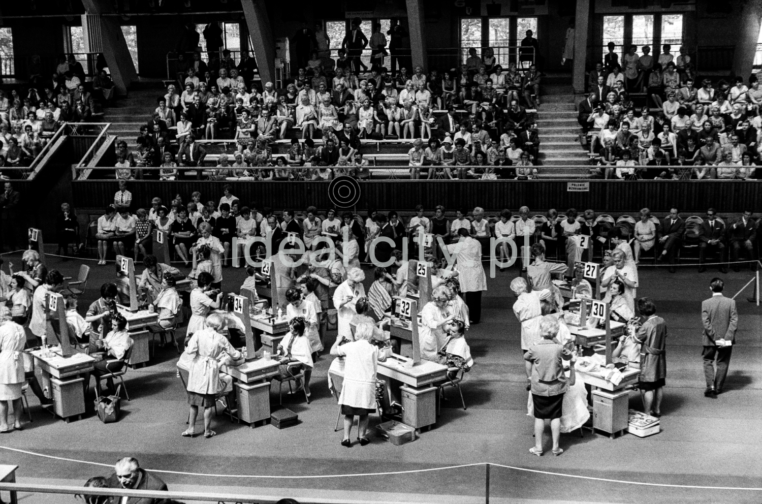 A shot from a height on the tables with participants set up in the center of the hall, and the audience in the background.