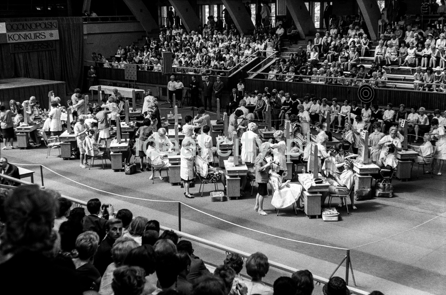 A shot from a height on the tables with participants set up in the center of the hall, and the audience in the background.