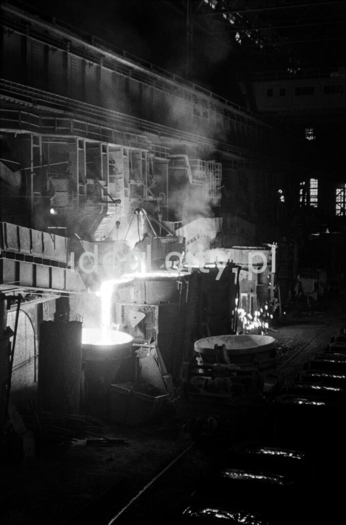 In a huge dark hall, molten iron is poured from one open vat to another.