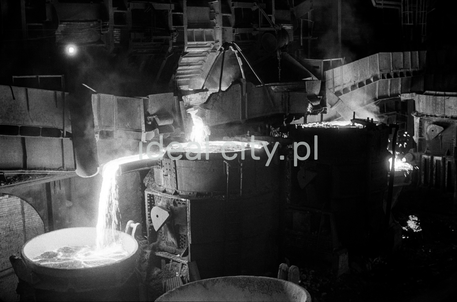 In a huge dark hall, molten iron is poured from one open vat to another.