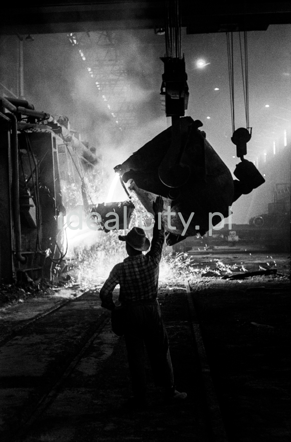 In a huge dark hall, a steelworker in a hat gives orders with his hand to the operator of a ladle suspended from the top, from which molten iron is poured from one open vat to another.