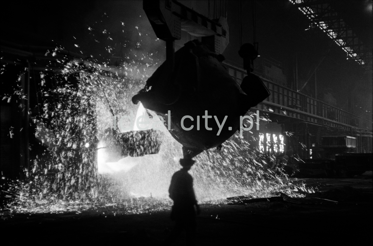 In a huge dark hall, a steelworker in a hat passes a vat, from which molten iron is poured into another.
