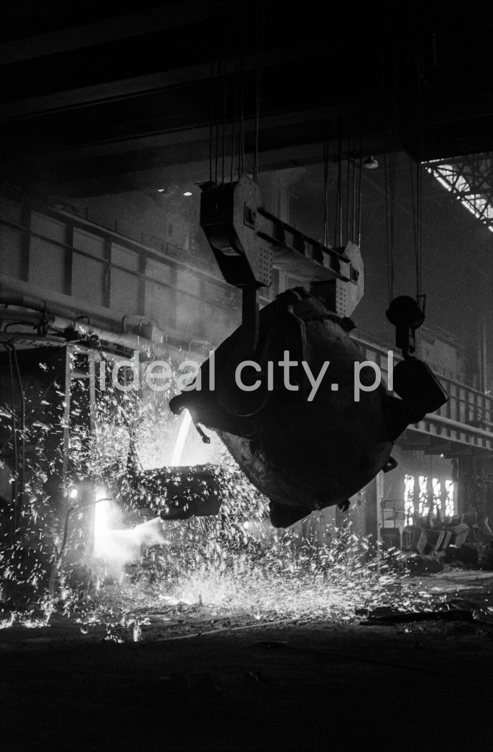 In a huge dark hall, molten iron is poured from one open vat to another.