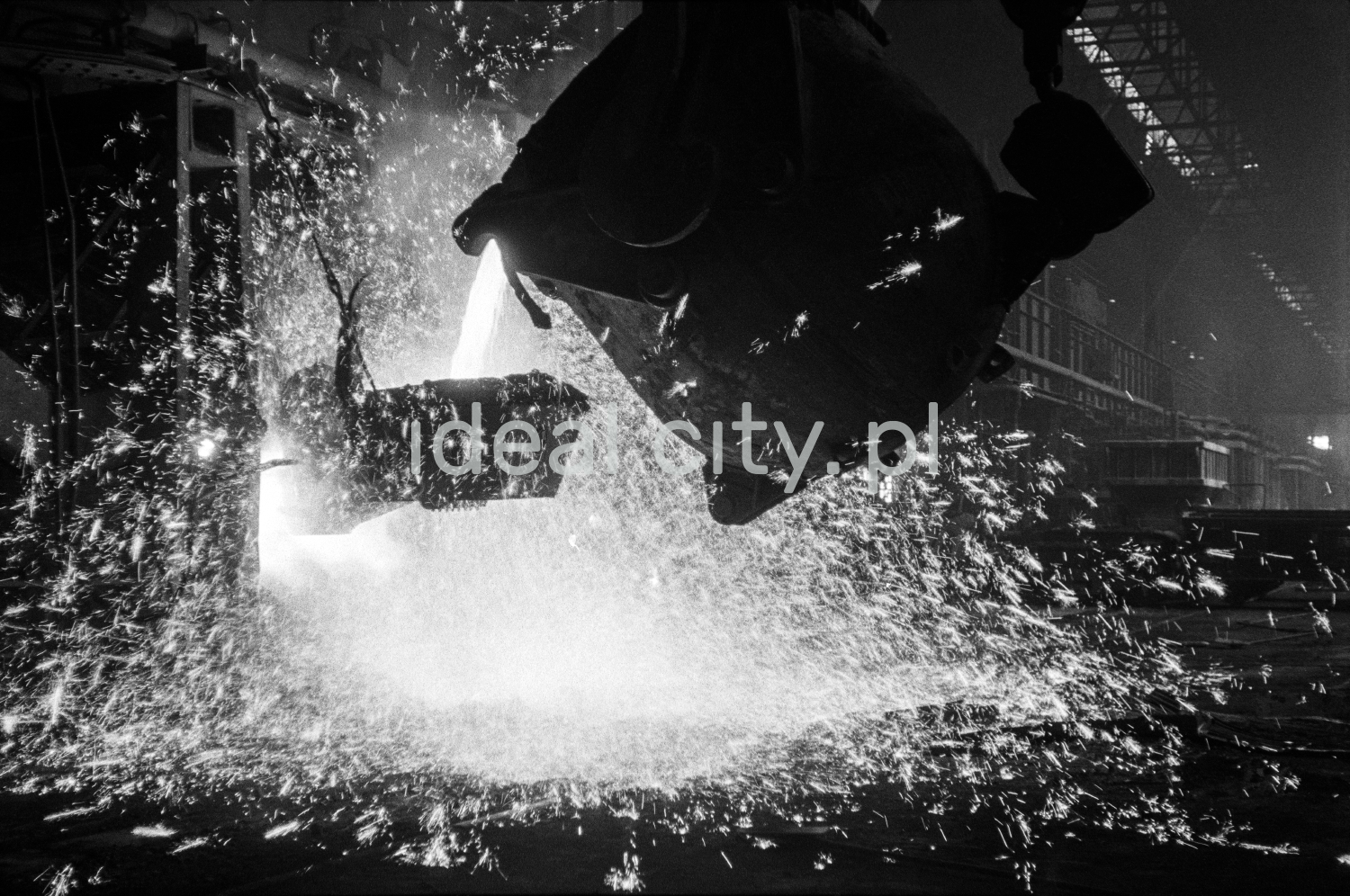 In a huge dark hall, molten iron is poured from one open vat to another.