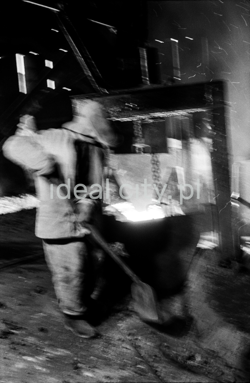 A steelworker in a full outfit cleans up the furnace surroundings with a shovel.