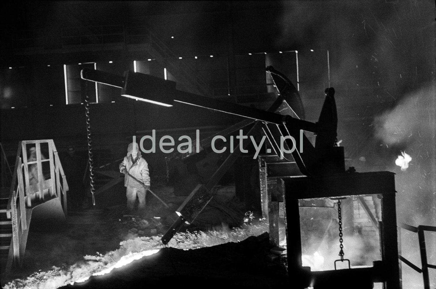 A steelworker in full outfit controls a stream of molten pig iron with a shovel.